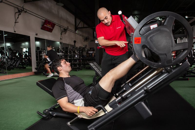 Un entrenador de Fit4Life le indica algo a un alumno en la prensa de piernas, con la sala de entrenamiento detrás