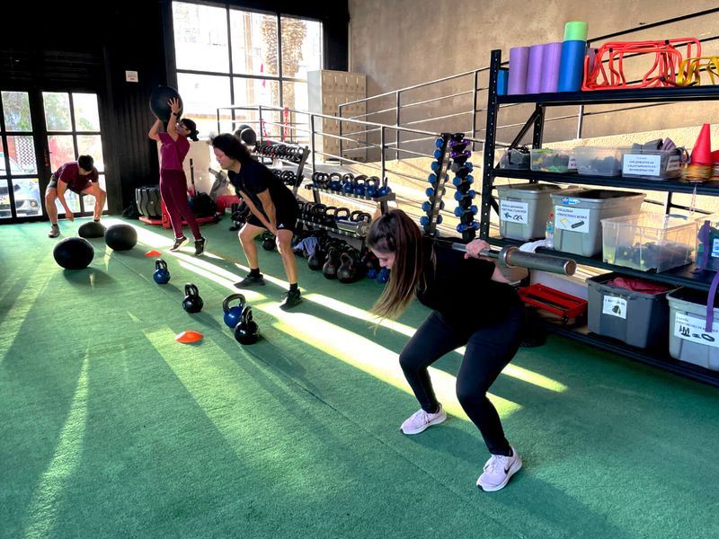 Una clase dirigida de Fit4Life: alumnos con balón, pesa rusa y barra sobre el pasto sintético